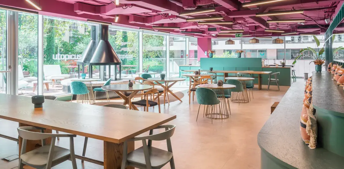 Un restaurante con mesas de madera y sillas verdes, ubicado junto a una ventana grande. En el techo, pintura rosa. Al fondo, un cuarto de estar al aire libre.