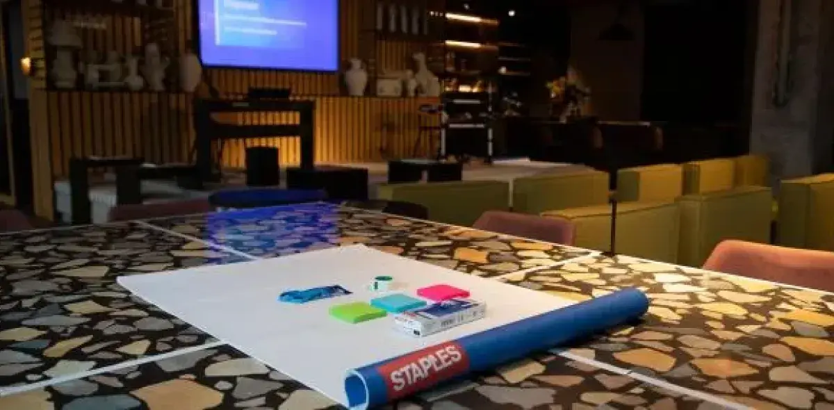 Papel y útiles de oficina sobre mesa con diseño de mosaico; pantalla de proyección y sillas en sala.