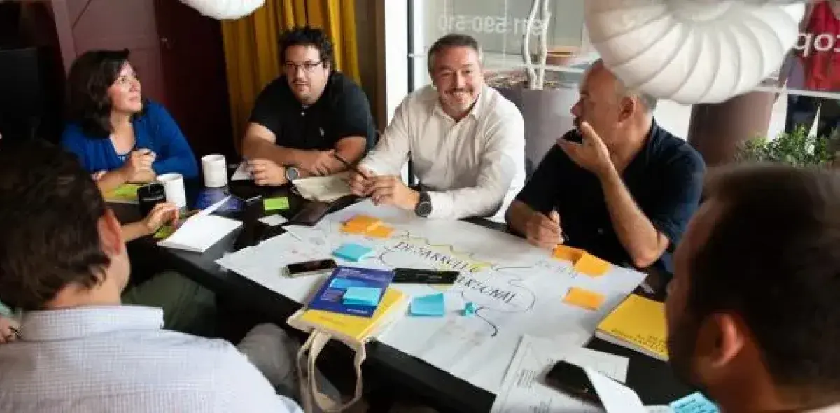 Personas sentadas conversando alrededor de una mesa, con papeles y notas adhesivas. La mesa está en un entorno interior, posiblemente una reunión de trabajo o taller colaborativo.
