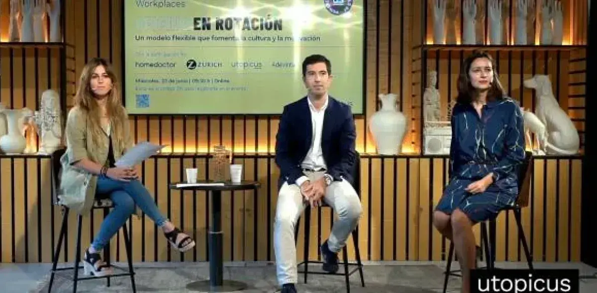 Tres personas sentadas en taburetes, participan en un panel. Detrás, un letrero dice: "EN ROTACIÓN Un modelo flexible que fomenta la cultura y la innovación". Ambiente decorado con jarrones blancos.