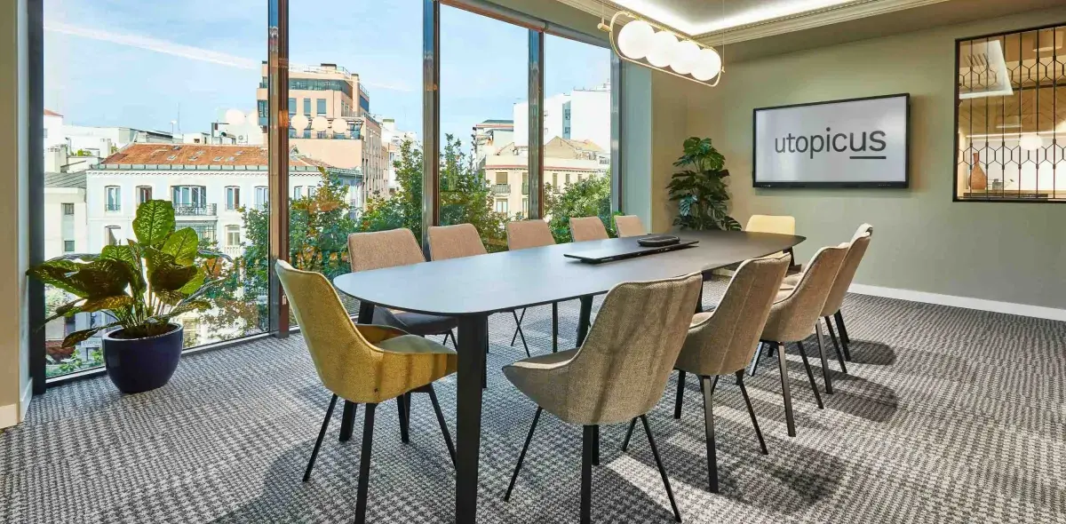 Una mesa de conferencias grande rodeada de sillas modernas se sitúa en una sala con ventanas grandes. En la pared, un monitor muestra el texto "utopicus".