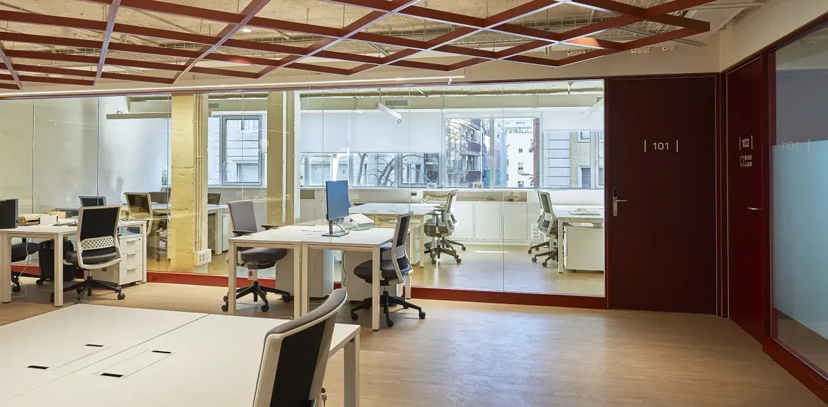 Oficina vacía con escritorios y sillas, rodeada de paneles de vidrio. En la puerta, numeración "101". Ambiente moderno con techos abiertos.