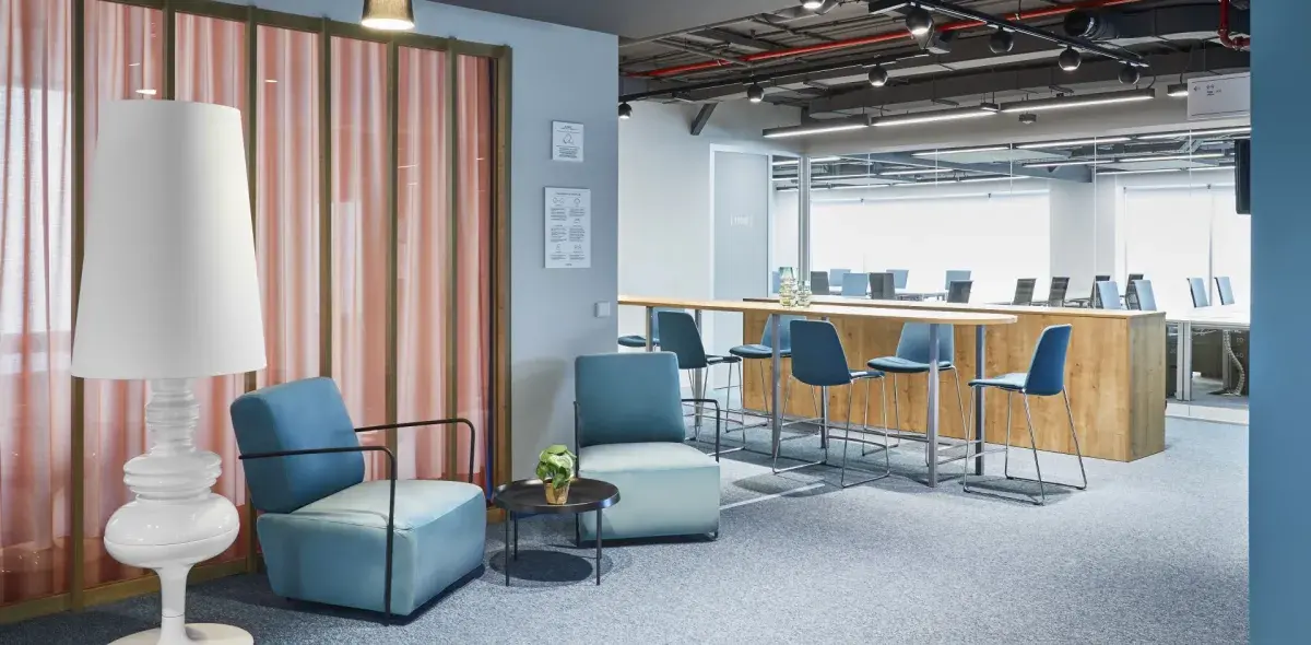 Una lámpara alta junto a sillones azules rodean una mesa pequeña. Al fondo, una barra de madera con sillas azules en un entorno de oficina moderna.
