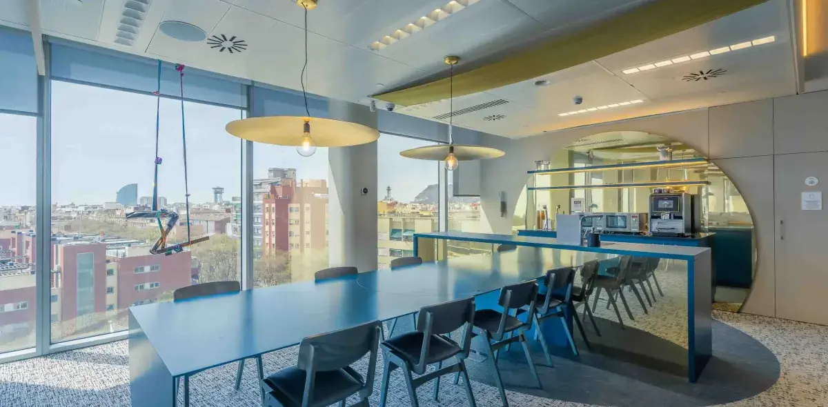 Mesa de comedor larga rodeada de sillas en una sala con amplias ventanas, lámparas colgantes y vista urbana. Una máquina de café y estanterías están al fondo.