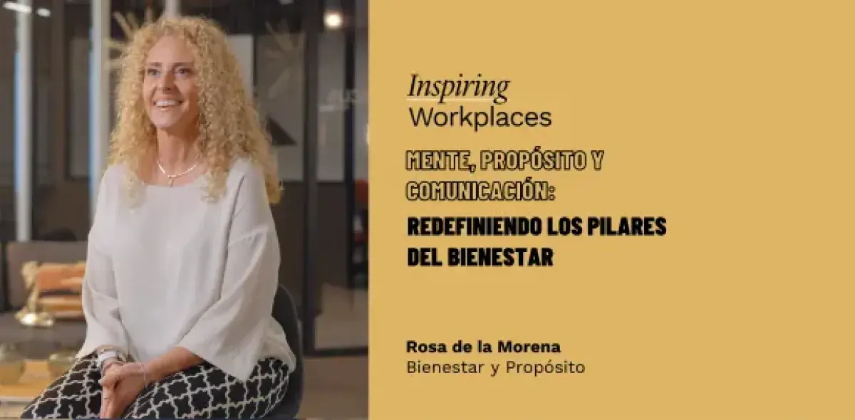 Mujer sentada sonriente, vistiendo blusa blanca y pantalones estampados en oficina moderna.