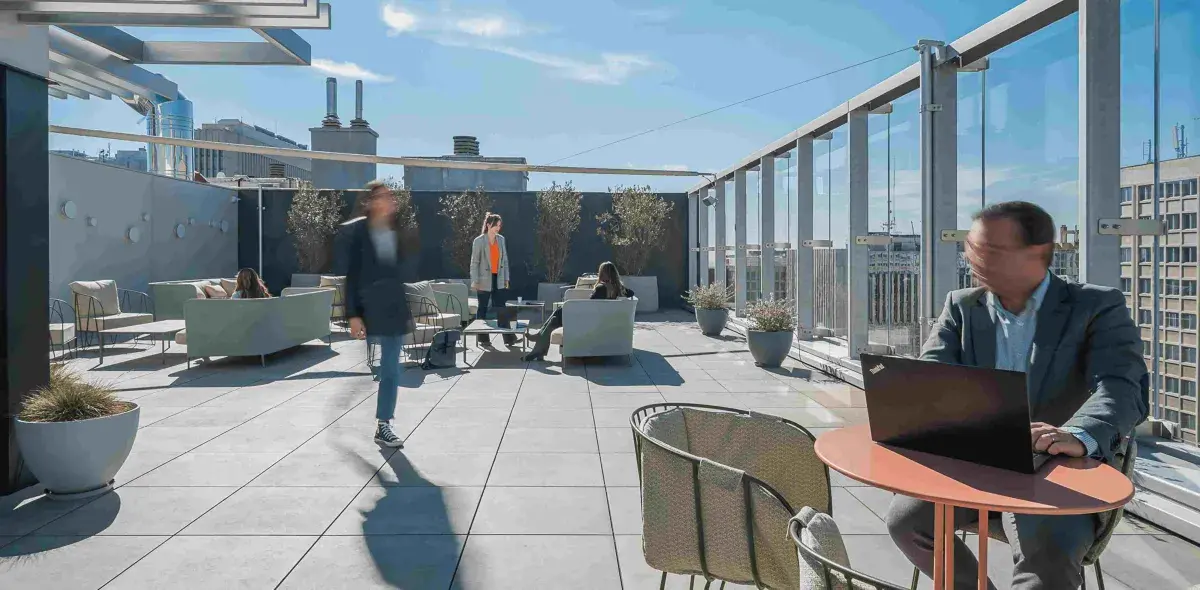 Persona sentada trabajando en una computadora portátil en una terraza soleada; otras personas socializan en sofás rodeados de plantas y edificios urbanos.