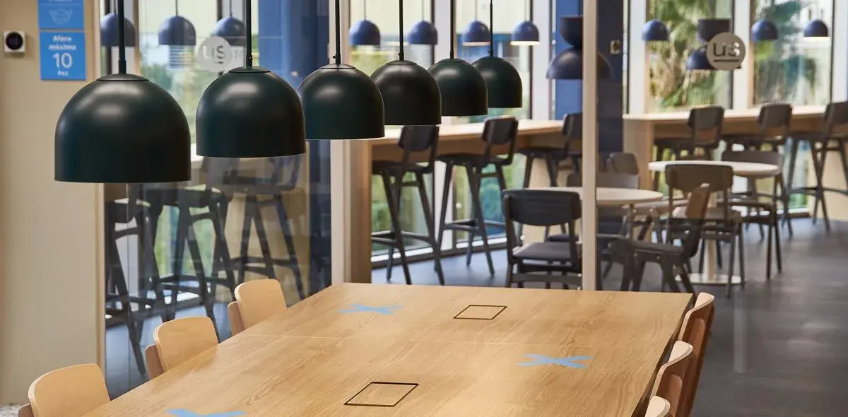 Mesa de madera clara con sillas alrededor, situada en comedor moderno; lámparas negras colgantes iluminan el espacio. En la pared, cartel azul: "aforo máximo 10 personas".