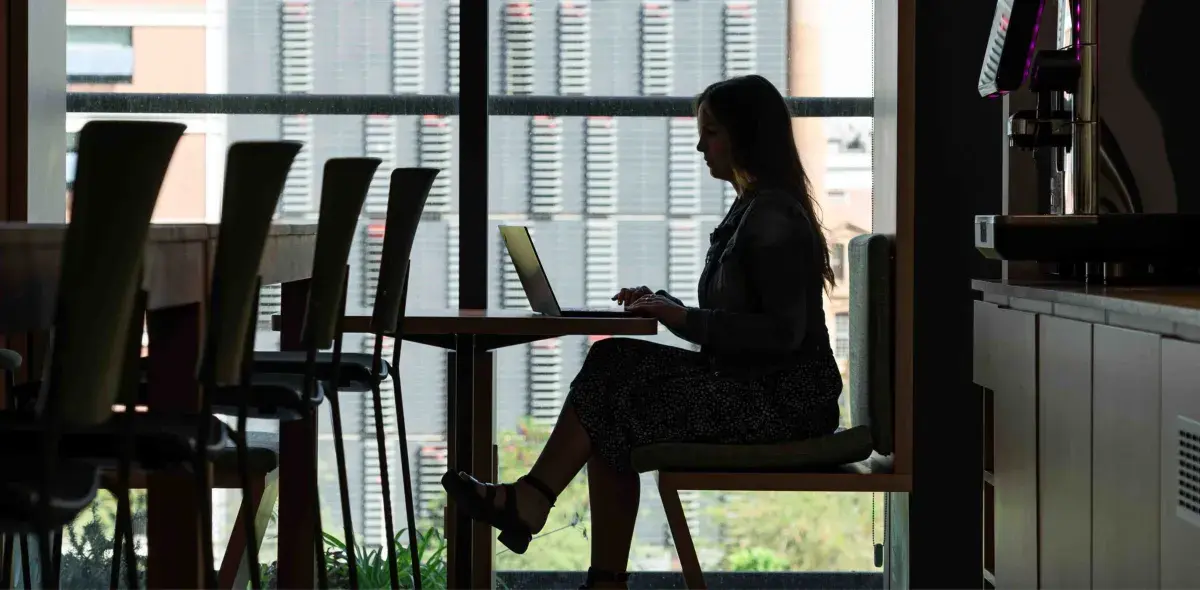 Persona trabajando en una computadora portátil, sentada en una mesa de cafetería con ventanales grandes al fondo.