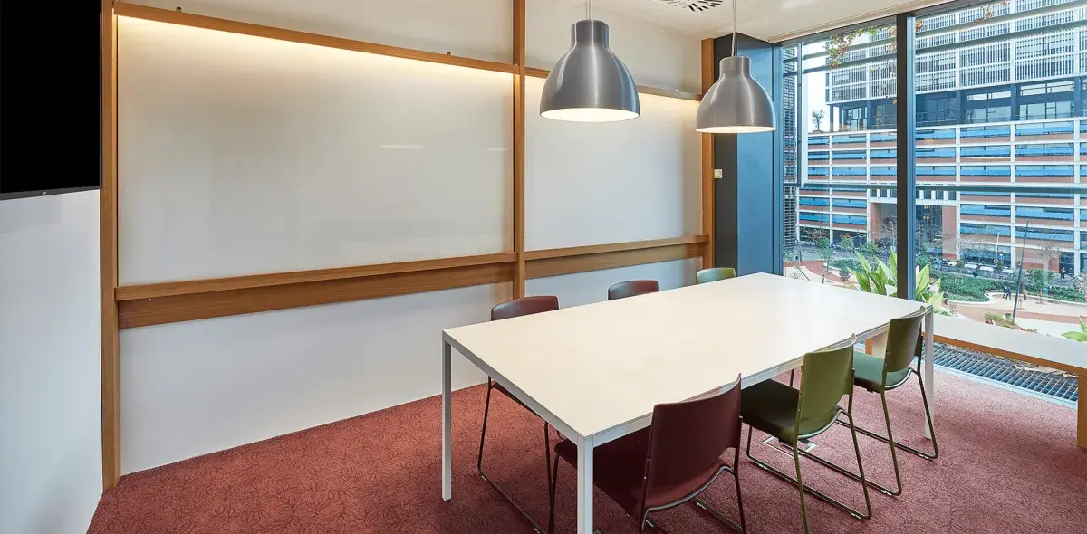 Una mesa blanca rectangular rodeada de sillas, bajo dos lámparas colgantes, en una sala de reuniones con ventana panorámica al exterior.