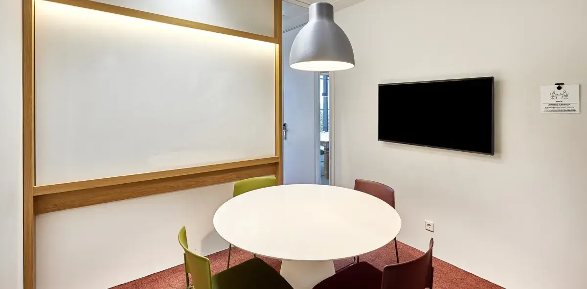 Mesa redonda rodeada de sillas de diferentes colores en una sala de reuniones con pantalla y pizarra iluminada. Lámpara colgante plateada sobre la mesa.