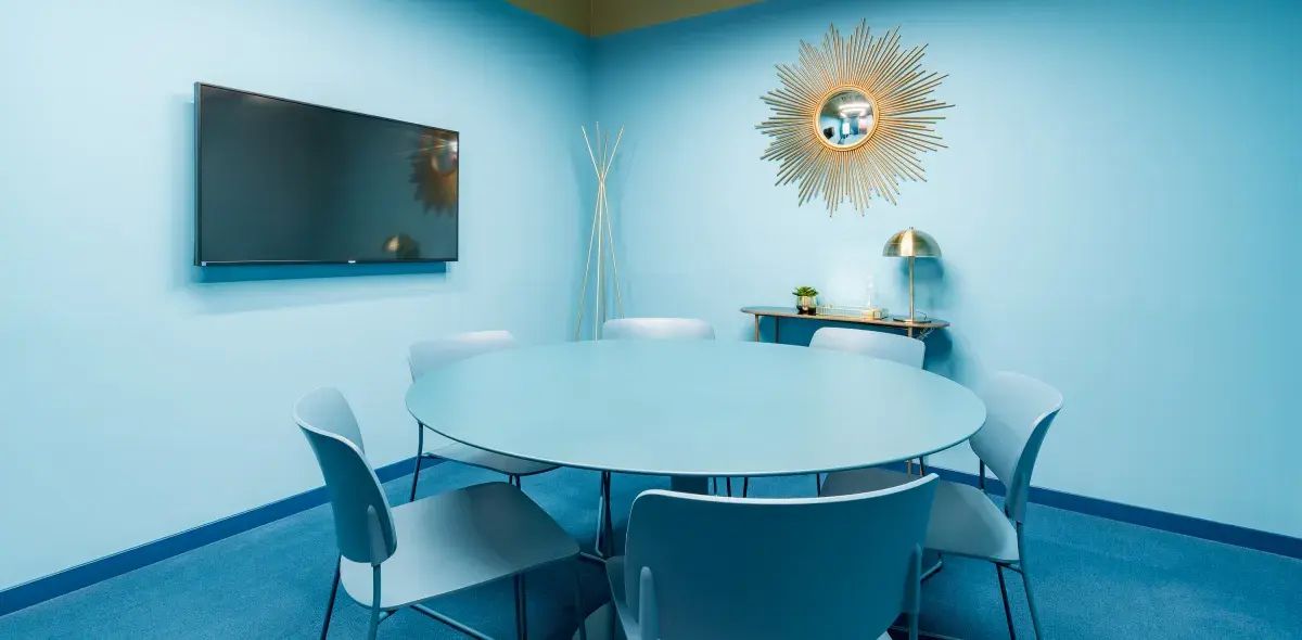 Mesa redonda rodeada de sillas, ubicada en una sala azul con televisor y espejo decorativo en la pared. Una lámpara y una pequeña planta adornan una consola al fondo.