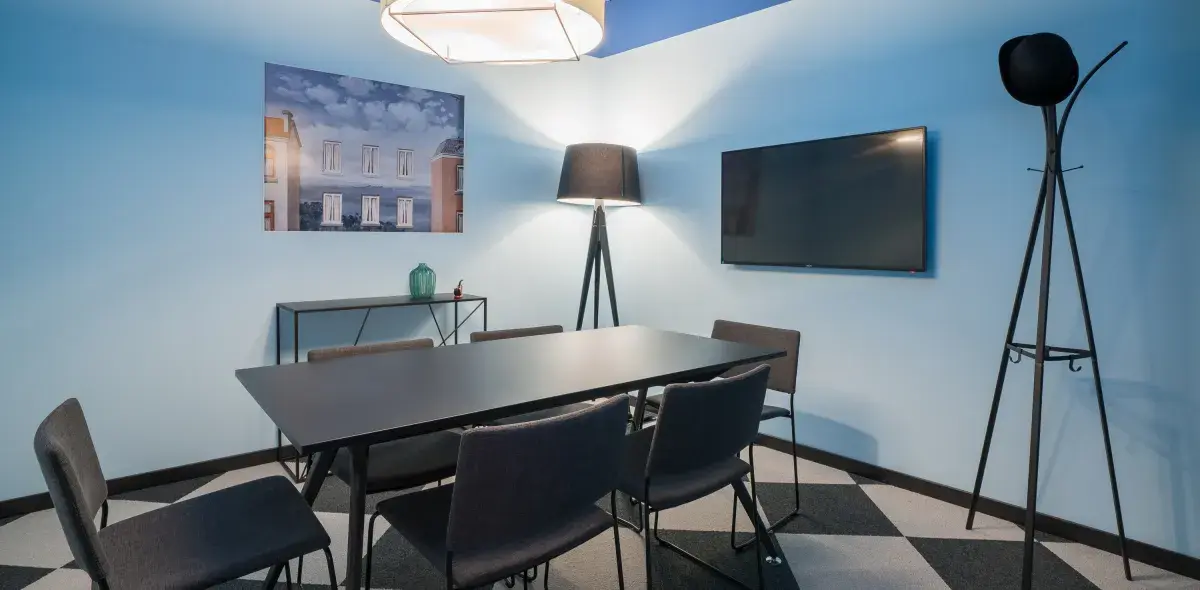 Una mesa rectangular negra con seis sillas rodea una lámpara de pie, en una sala con paredes azules y televisor montado. Una pintura de edificios decora la pared.