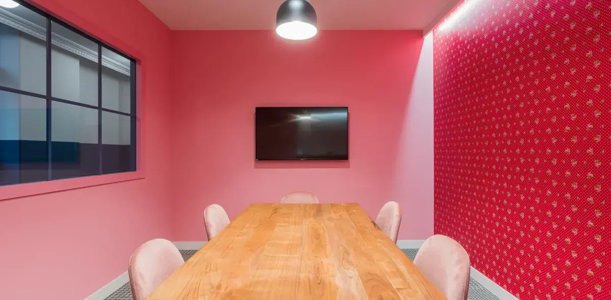 Una mesa de madera se sitúa bajo una lámpara colgante en una sala de paredes rosadas, con silla alrededor y un televisor en la pared.