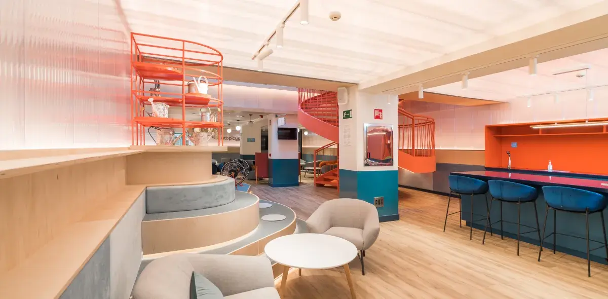 Estantería naranja con objetos decorativos, situada en un espacio interior moderno con sillas grises, mesas blancas y escaleras en espiral. Al fondo, letrero que dice "topicus".