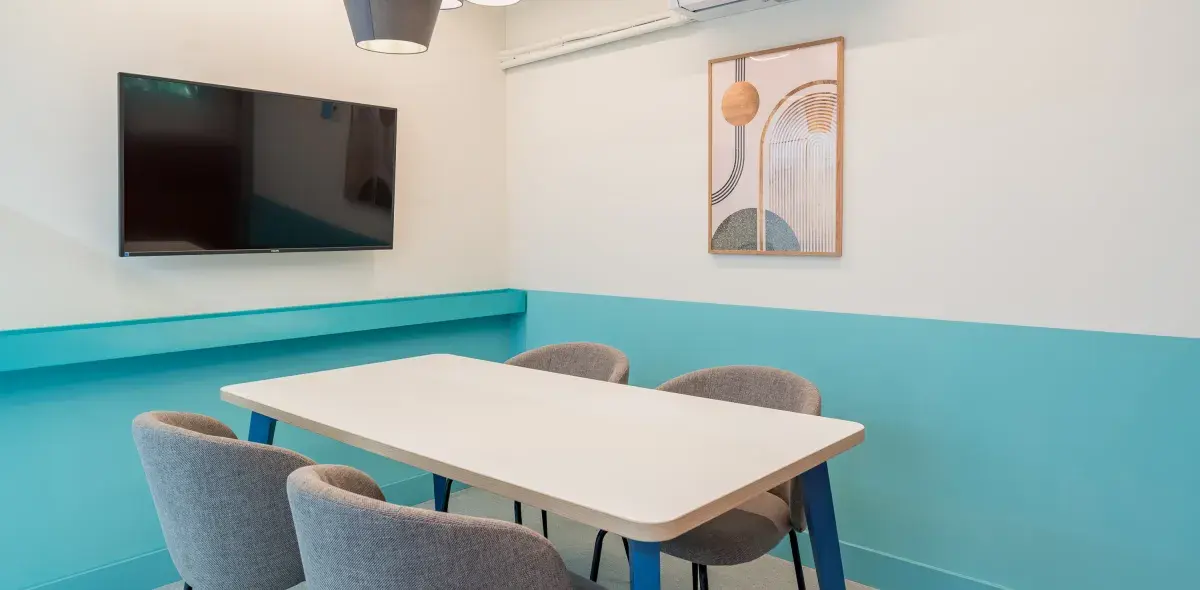 Una mesa blanca con cuatro sillas grises está en una sala con paredes azul y blanca. Un televisor está montado en la pared, junto a un cuadro abstracto.