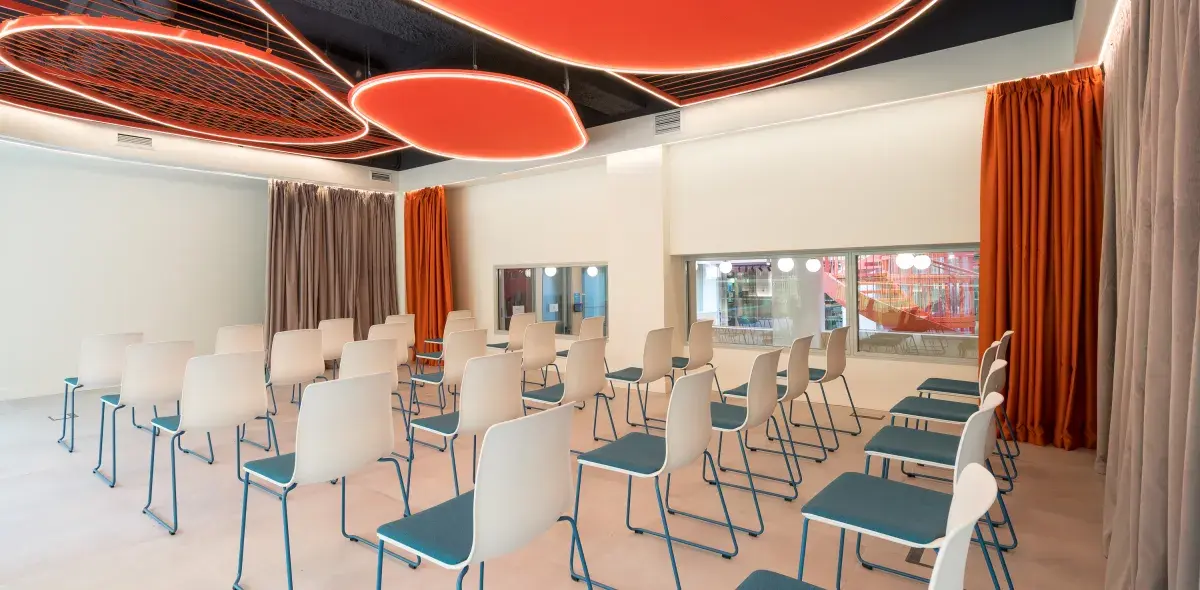 Sillas de respaldo blanco y asiento azul están ordenadas en filas; techo con círculos naranjas iluminados y cortinas a los lados en una sala moderna.