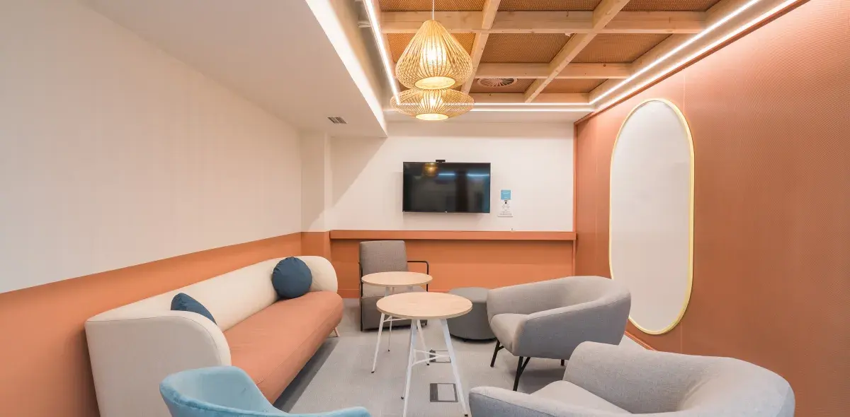 Sofá moderno y sillas grises rodean mesas, en sala de espera con pared naranja y televisor montado. Decoración luminosa y espejo ovalado en la pared derecha.