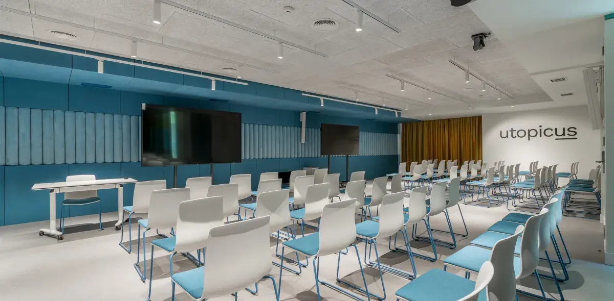 Filas de sillas blancas y azules enfrentan pantallas negras en una sala de conferencias moderna.