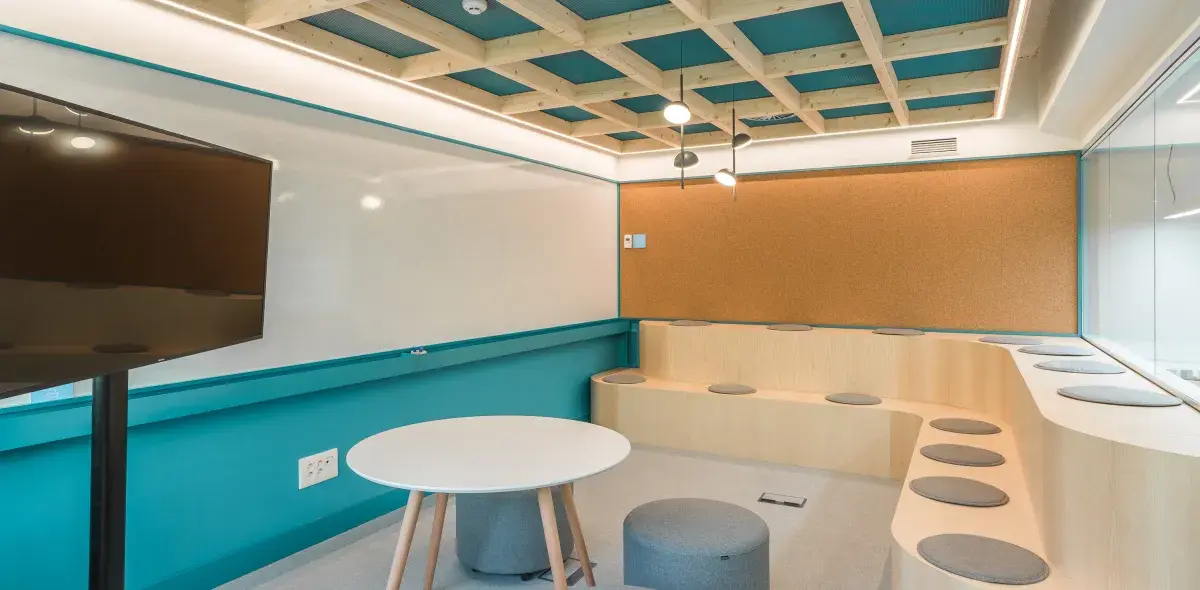 Pantalla negra fija al lado de una pizarra en sala moderna con techos de madera. Mesas redondas y asientos grises complementan el entorno.
