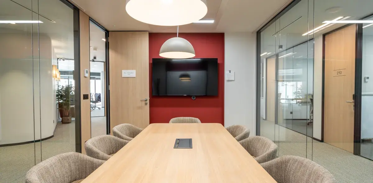 Mesa rectangular rodeada de sillas sin personas, ubicada en una sala de reuniones con paredes de vidrio y una pantalla grande en un muro rojo. Puerta con texto: "Main Negotiation Room".