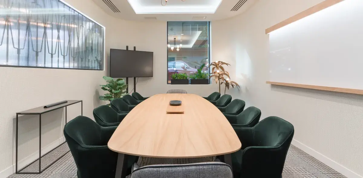 Mesa larga de madera rodeada por sillas en una sala de reuniones con televisor, plantas y una pizarra. Ventana al fondo deja ver un auto afuera.