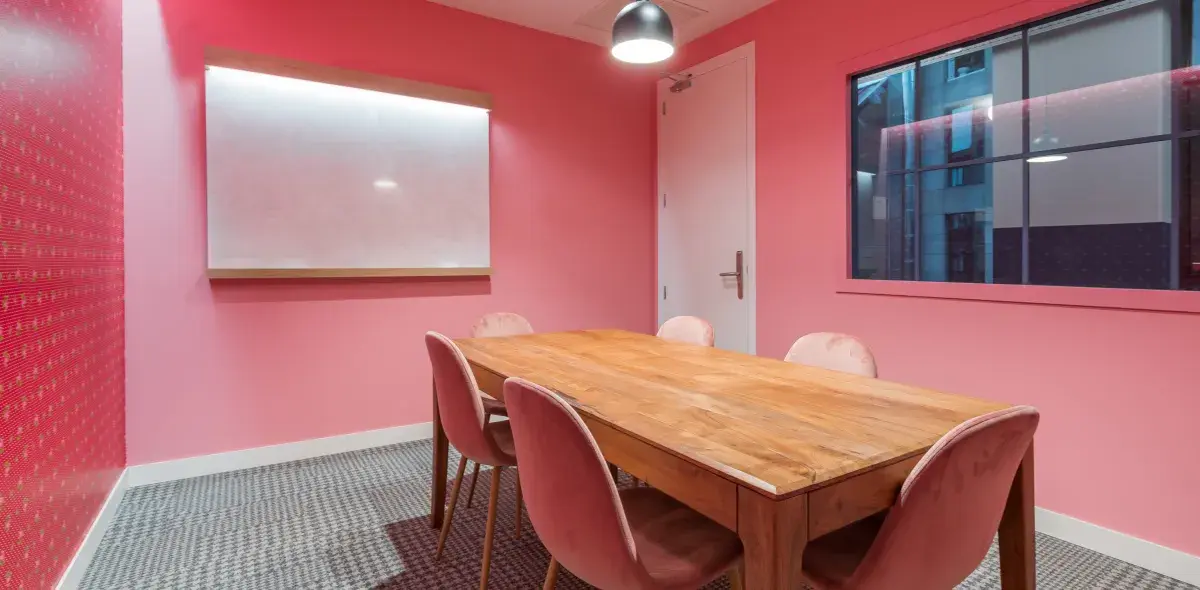 Mesa de madera rodeada por seis sillas rosas. En la pared, pizarra blanca iluminada. Habitación con paredes rosadas, lámpara colgante y ventana al exterior.