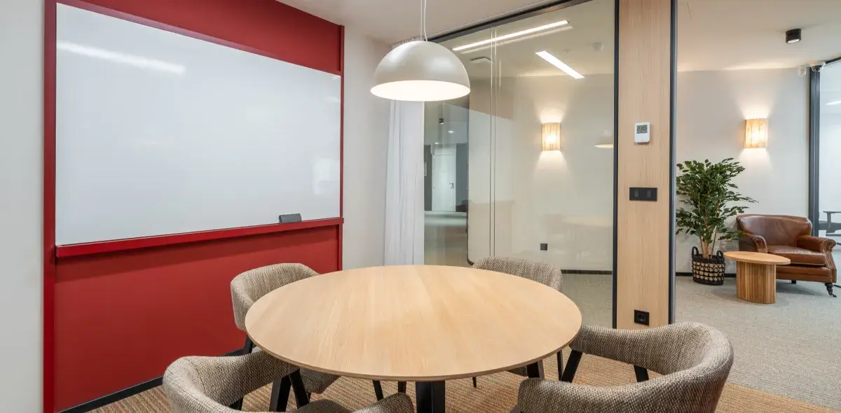 Una mesa redonda de madera rodeada de sillas en una sala de reuniones con pizarra y paredes de cristal. Hay un sillón marrón y una planta al fondo.
