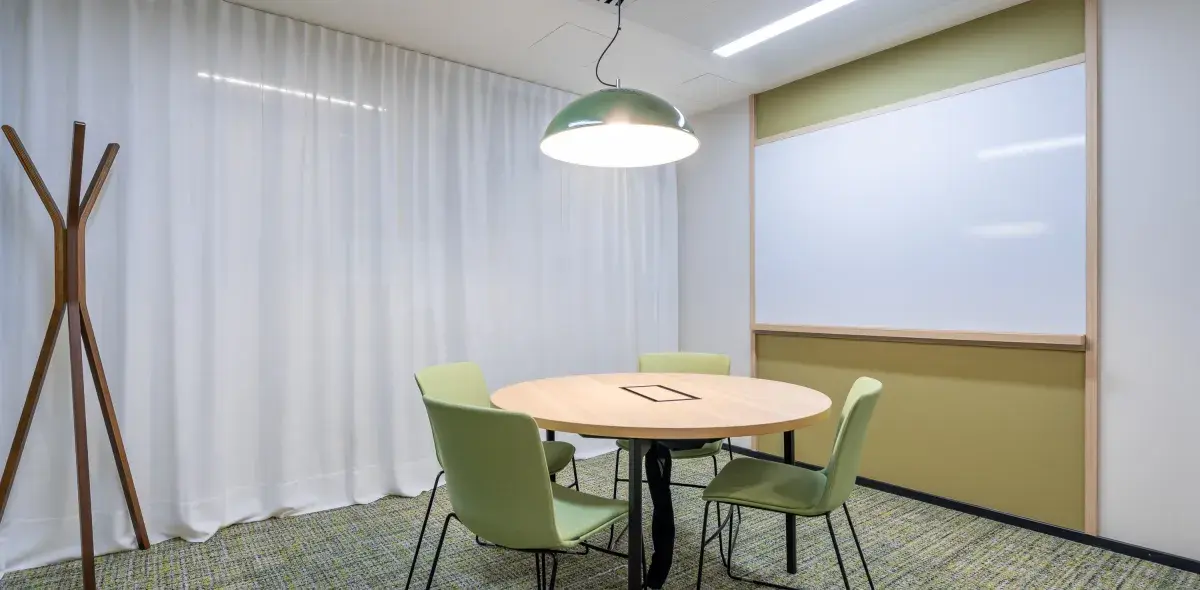 Mesa redonda rodeada de sillas verdes estática en una sala de reuniones con cortinas blancas y pizarra blanca. Un perchero de madera se sitúa a la izquierda.