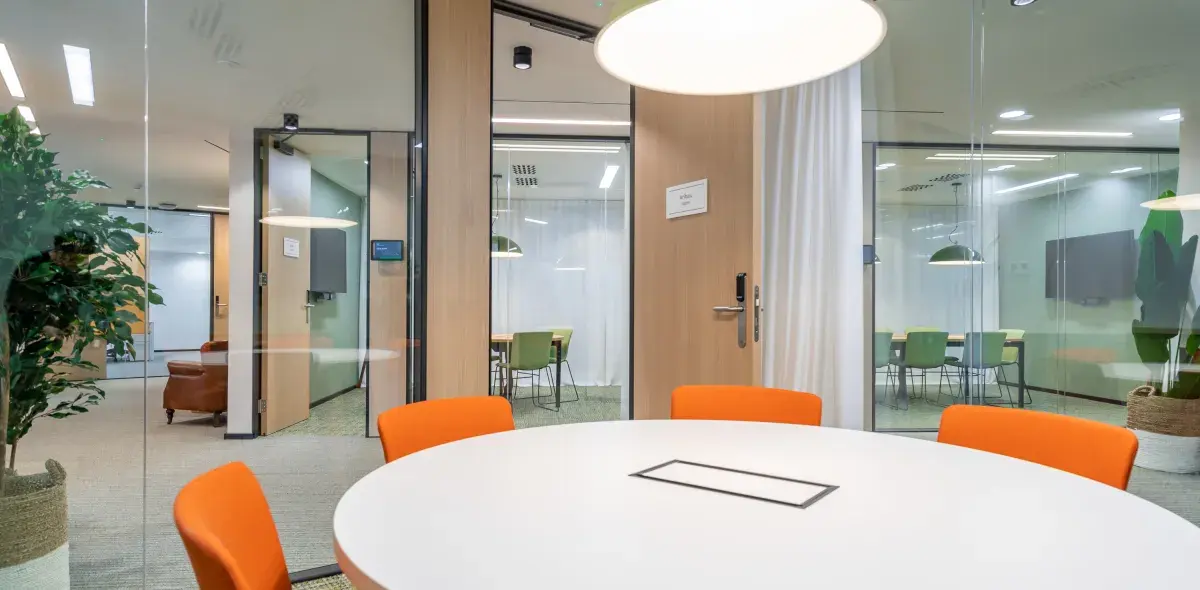 Una mesa redonda blanca con sillas naranjas está en una sala de reuniones moderna con puertas de vidrio. Al fondo, se ven más mesas, sillas verdes y plantas decorativas.