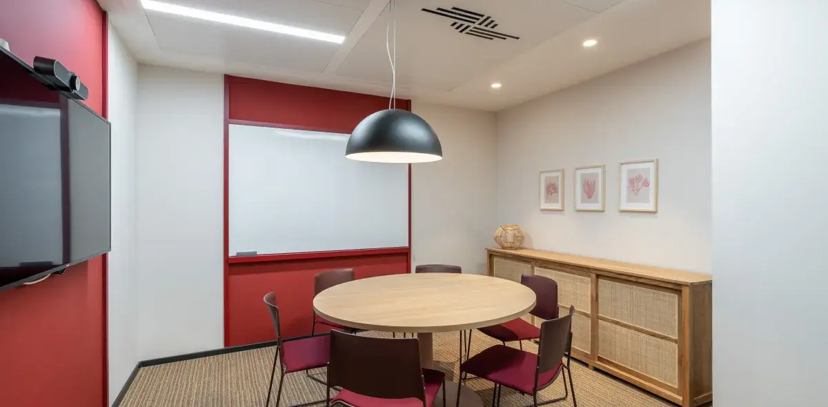 Una mesa redonda con sillas rodea una lámpara colgante en una sala de reuniones pequeña, con pizarra, televisor y muebles decorativos.