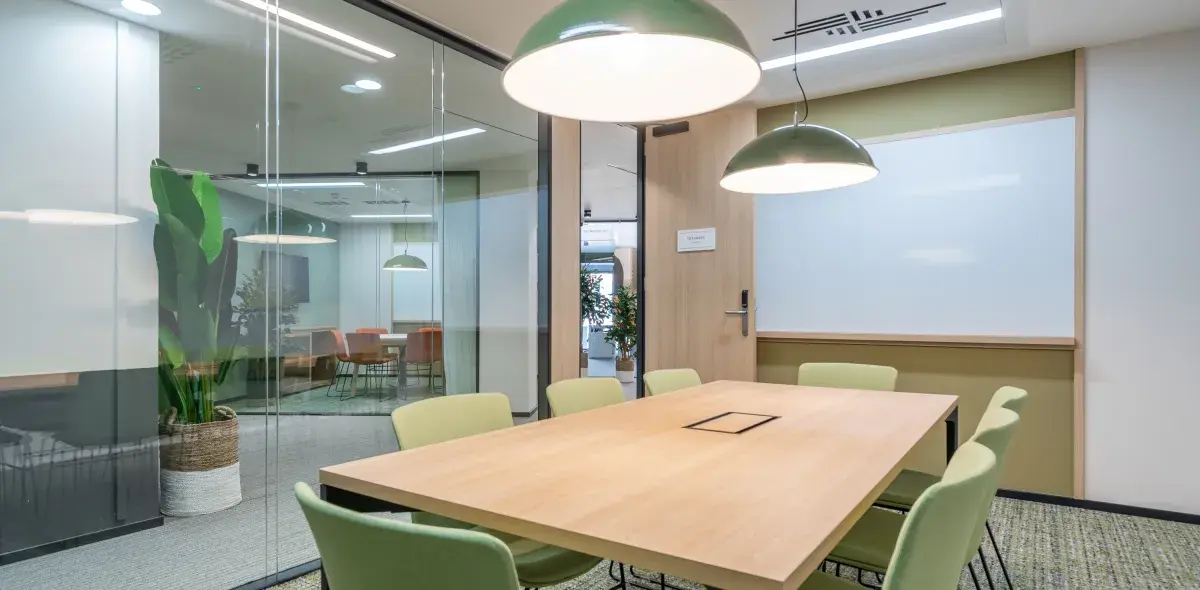 Mesa de madera rodeada por sillas verdes en sala de reuniones. Lámparas colgantes alumbran, planta decorativa al fondo. Puerta con cartel que dice "Colabora".