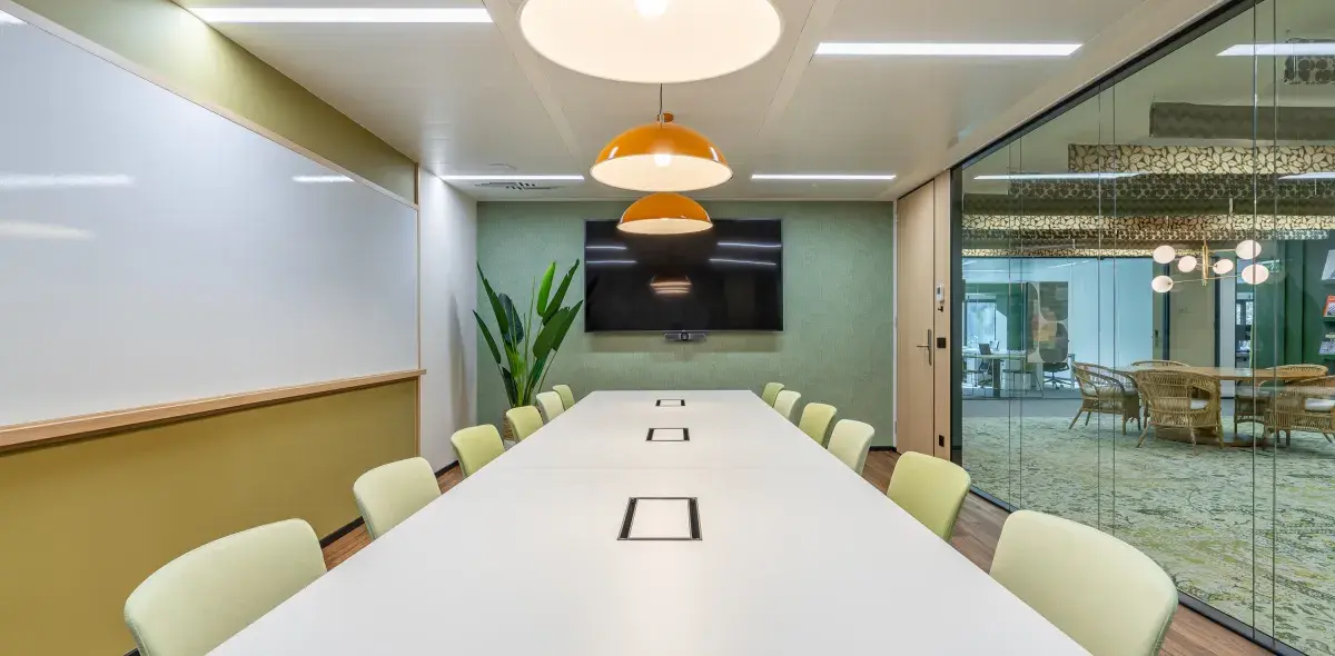 Mesa larga blanca en sala de conferencias, rodeada de sillas, con pantalla en pared verde. Lámparas colgantes naranjas iluminan. Pizarra blanca a un lado, planta decorativa cercana.