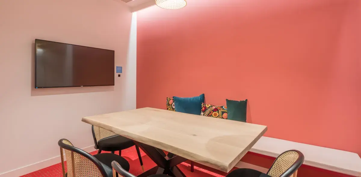 Mesa grande de madera con sillas alrededor, situada en una sala con pared rosa. Televisión colgada a la izquierda. Cojines decorativos en un banco contra la pared.