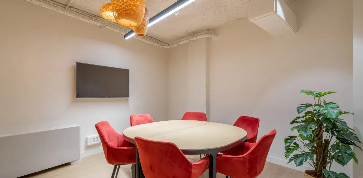 Mesa redonda rodeada de sillas rojas, situada en una sala con televisor montado en la pared, lámpara colgante y planta decorativa.