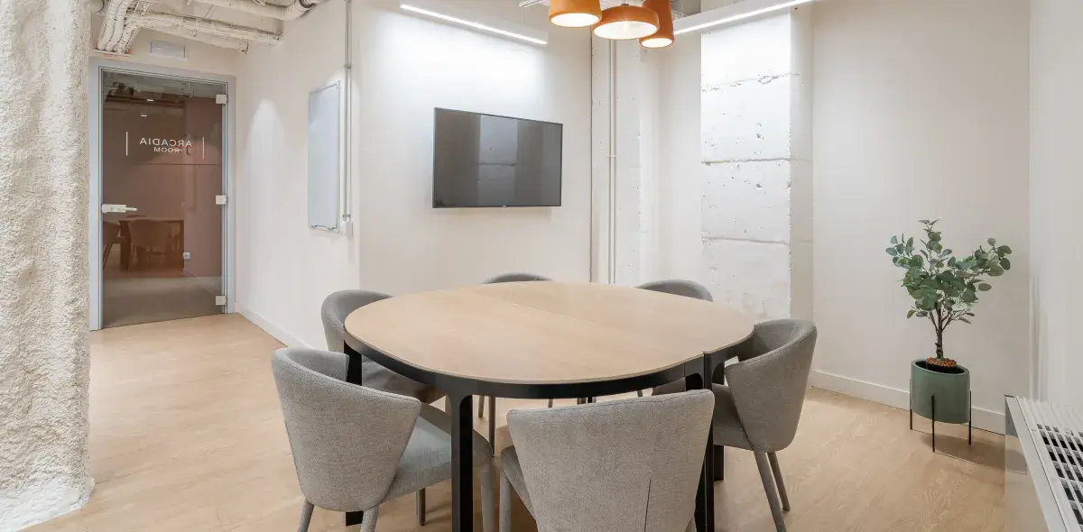 Mesa redonda rodeada de sillas grises estática en una sala de reuniones moderna con televisor y planta decorativa. En la puerta se lee "Arcadia Room".
