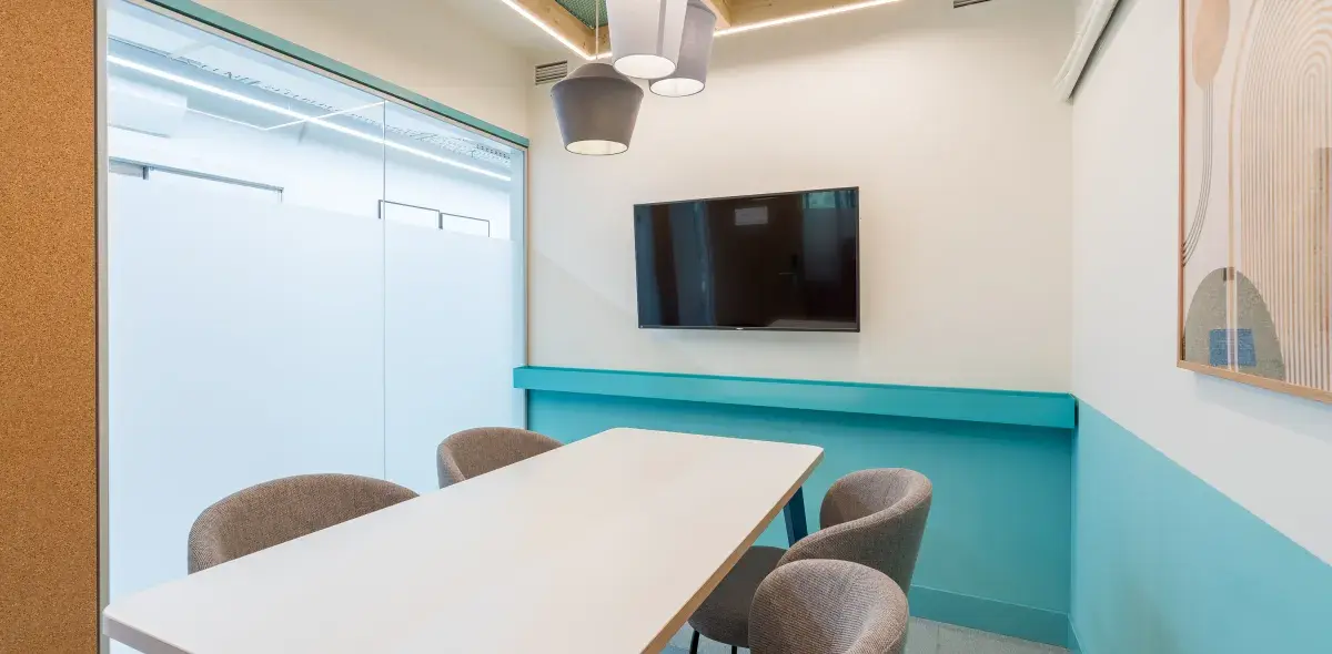 Mesa blanca con cuatro sillas grises en una sala moderna; pantalla negra colgada en la pared y lámparas colgantes grises iluminan el espacio. Paredes con diseño minimalista.