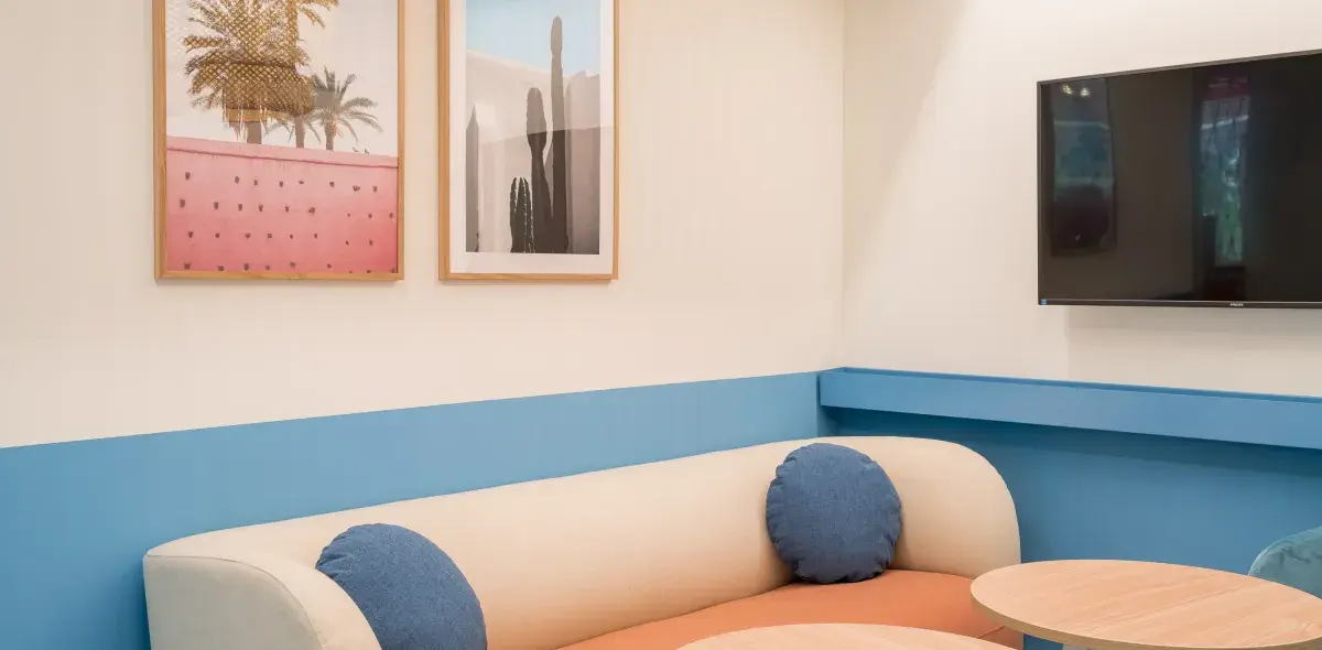 Sofá beige con cojines azules ocupa una esquina de sala, decorada con cuadros y televisor.