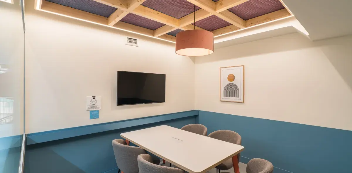 Una mesa blanca rodeada de sillas grises se encuentra en una sala con televisión en la pared y un cuadro decorativo. Luz cálida cuelga del techo de madera.
