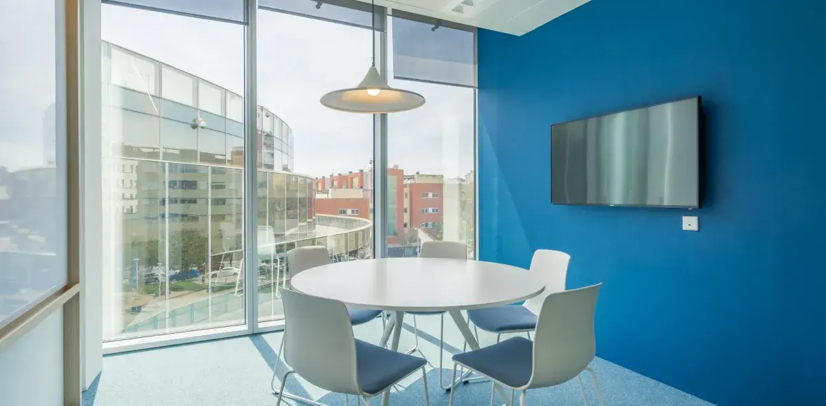 Mesa redonda con sillas blancas y grises, situada en una sala de reuniones con paredes azules y una gran ventana. Una televisión está montada en la pared.