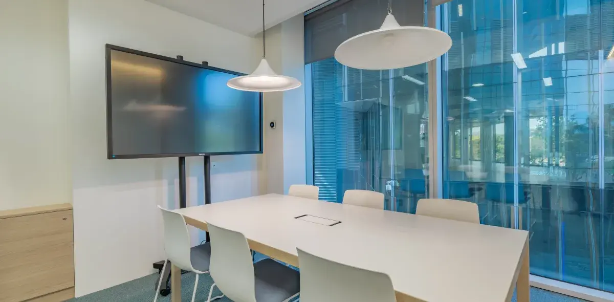Una sala de reuniones con una mesa blanca, ocho sillas, dos lámparas colgantes y una gran pantalla en la pared; ventanales al fondo.