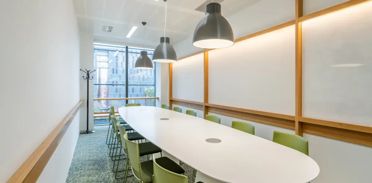 Mesa ovalada blanca rodeada de sillas verdes, iluminada por lámparas colgantes. Ubicada en una sala de reuniones con paredes blancas y ventanas grandes.