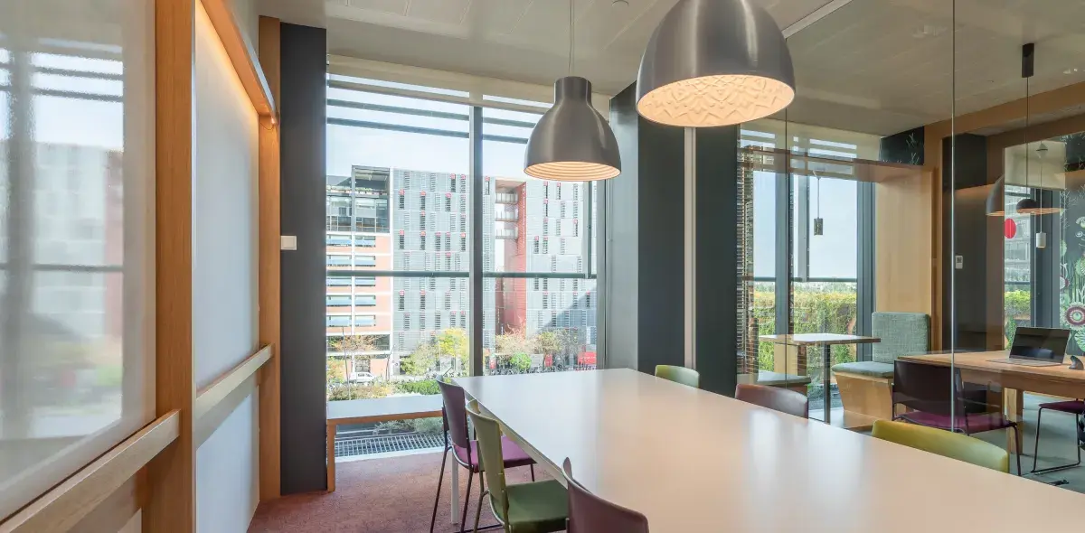 Una mesa alargada con sillas de colores, iluminada por lámparas colgantes, en una sala de reuniones con ventanales y vistas a un edificio moderno exterior.