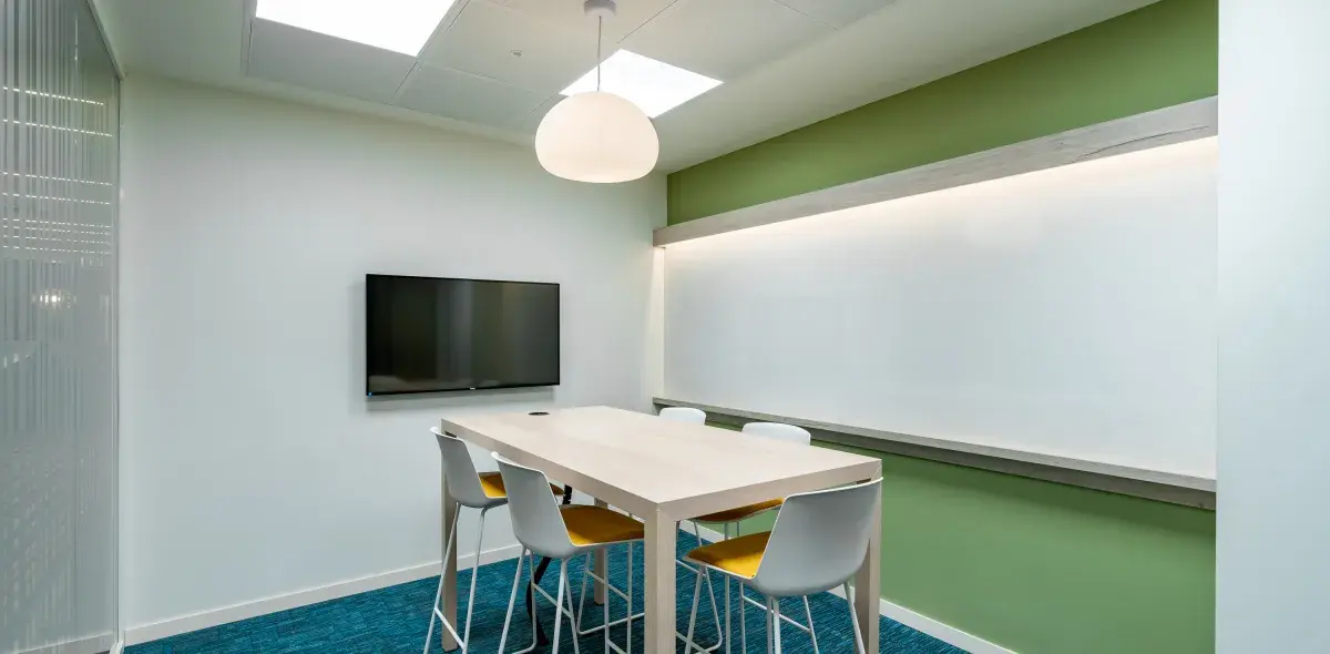 Una mesa alta con sillas modernas está en una sala de reuniones pequeña. Hay una pantalla de televisión montada en la pared blanca y una pizarra al fondo con pared verde. Unas luces del techo iluminan el espacio con alfombra azul.