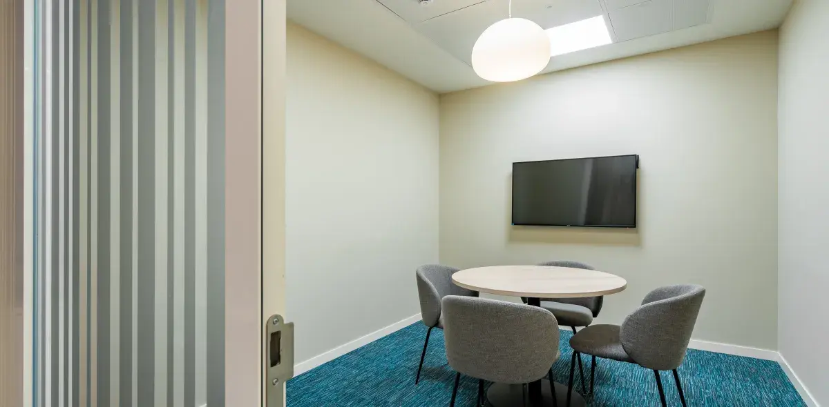 Mesa redonda rodeada de cuatro sillas grises, ubicada en una sala pequeña con alfombra azul, televisor en la pared y lámpara colgante blanca. Puerta corredera semiabierta.