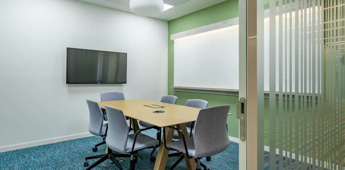 Una mesa de conferencias con seis sillas rodea un monitor en una sala de reuniones con paredes verdes y alfombra azul. Una puerta de vidrio está parcialmente abierta.