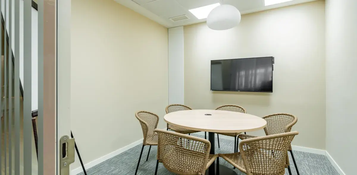Una mesa ovalada rodeada de sillas tejidas está en una sala de reuniones con televisión montada en la pared. La iluminación es suave y el piso es alfombrado.