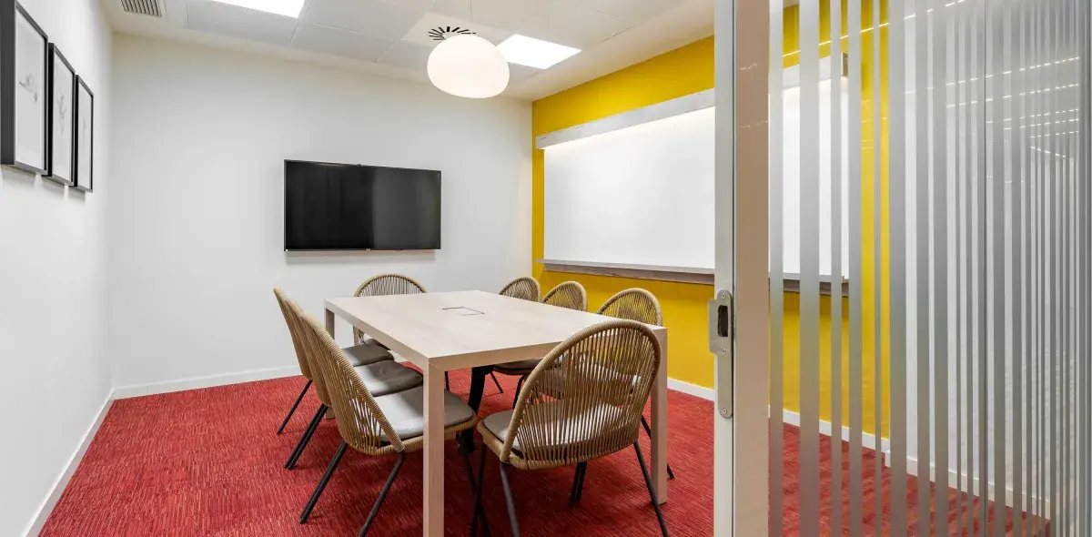 Una mesa rectangular rodeada de seis sillas está en una sala con paredes blancas y amarillas, una pared tiene un televisor y otra un tablero blanco.