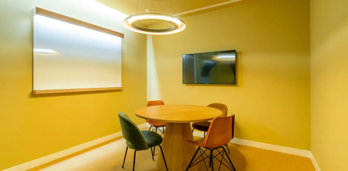 Una mesa redonda rodeada de sillas se encuentra en una sala de reuniones, con una pantalla y una pizarra blanca en las paredes amarillas.