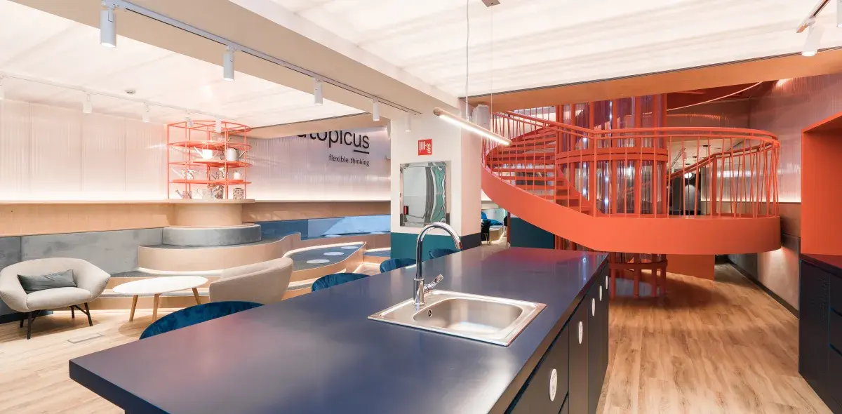 Isla de cocina con fregadero en primer plano; al fondo, escalera en espiral roja y sala con sofás.