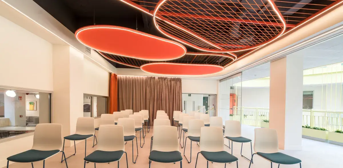 Sillas blancas alineadas están en una sala de conferencias moderna, con techos decorativos y cortinas marrones.